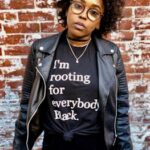 Home Confident Black woman in a leather jacket and statement tee against a brick wall.